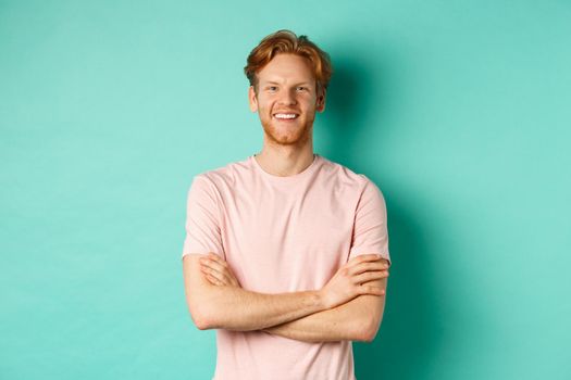 Portrait of friendly-looking young man with red hair and beard smiling and starng satisfied, holding hands crossed on chest, standing over turquoise background.