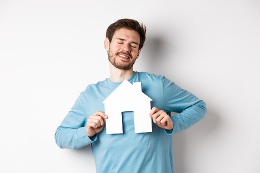 Real estate and insurance concept. Dreamy young man smiling with closed eyes, showing paper house cutout, wishing to buy home, standing over white background.