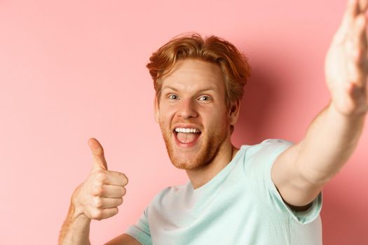 Excited redhead man tourist taking selfie and showing thumbs-up, holding camera with stretched out hand, standing over pink background.