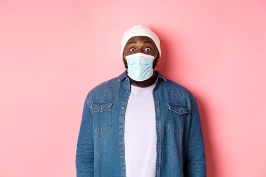 Covid-19, lifestyle and quarantine concept. Image of impressed african-american man in face mask staring at camera, gasping surprised, standing over pink background.