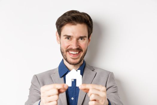 Real estate and insurance concept. Handsome man in suit smiling, showing small paper house cutout, standing on white background.