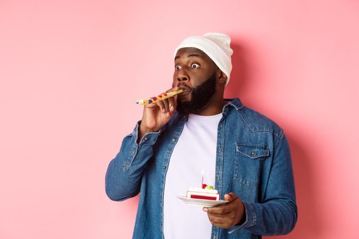 Happy Black hipster celebrating birthday, blowing party whistle, holding bday cake with candle, standing over pink background.