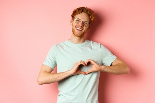 Valentines day concept. Handsome redhead man in glasses, showing heart sign and say I love you, standing over pink background.