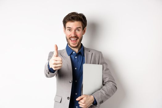 Good job. Happy smiling business man in suit holding laptop, showing thumb up to praise you, nice work gesture, standing on white background.