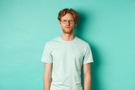 Confused young redhead man in glasses staring at camera puzzled, squinting perplexed, cant understand something, standing over mint background.