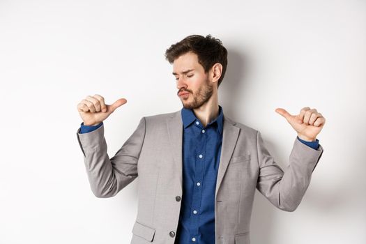 Handsome and confident man in suit pointing at himself, feeling cool and sassy, self-promoting his good appearance, standing on white background.