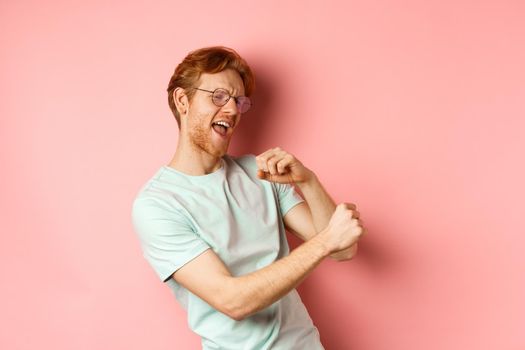 Summer vacation concept. Happy young man with red hair dancing and having fun, singing along music, standing over pink background.