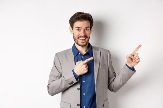 Happy businessman in suit pointing fingers right and smiling excited, showing special deal, standing on white background.