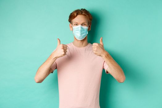 Covid-19, pandemic and lifestyle concept. Cheerful redhead guy in medical mask showing thumbs up in approval, like and praise product, standing over turquoise background.