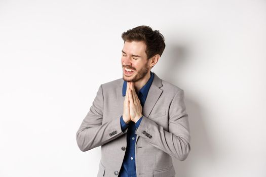 Happy and thankful businessman rejoicing, holding hands in begging or namaste sign, thanking for help, smiling relieved, standing in suit on white background.