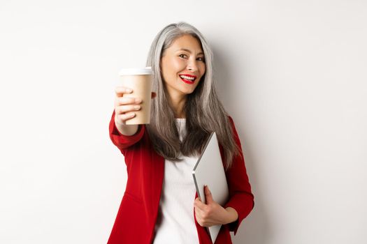 Business. Cheerful asian woman manager giving you cup of coffee and smiling, standing with laptop in hand, white background.