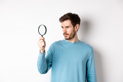 Young handsome man look through magnifying glass with curious face, investigating or searching for something, standing on white background.