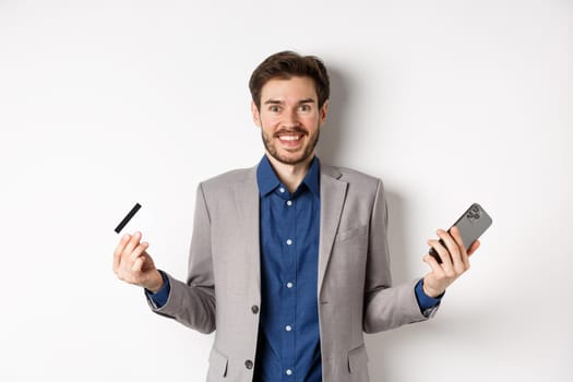 Online shopping. Excited man making money, smiling amazed, holding smartphone and credit card, standing against white background in suit.