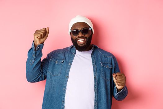 Happy Black man in beanie an sunglasses rejoicing, dancing with happy face, standing over pink background.