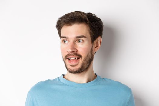 Close-up of surprised and excited bearded man, looking left with opened mouth and amazed face, checking out promo, white background.
