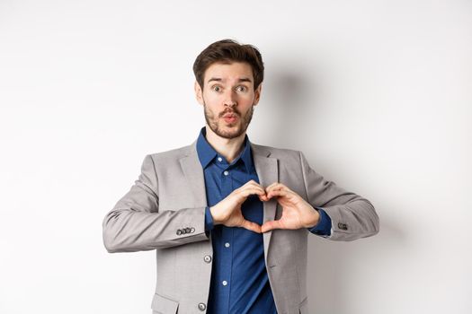 Handsome man looking silly, showing heart love you sign and pucker lips, waiting for kiss from lover, standing in suit on white background.