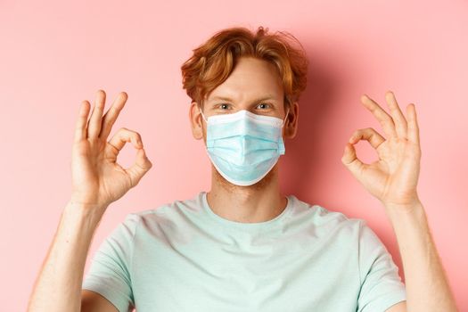 Covid-19 and pandemic concept. Handsome guy with messy ginger hair, wearing medical mask on face and showing okay signs, standing over pink background.