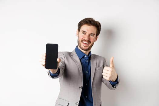 E-commerce and online shopping concept. Cheerful businessman in suit showing thumbs up and empty smartphone screen, recommending app, standing on white background.