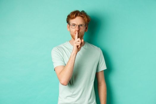Serious redhead man hushing, tell to be quiet, making shush gesture and looking at camera, standing in t-shirt over turquoise background.