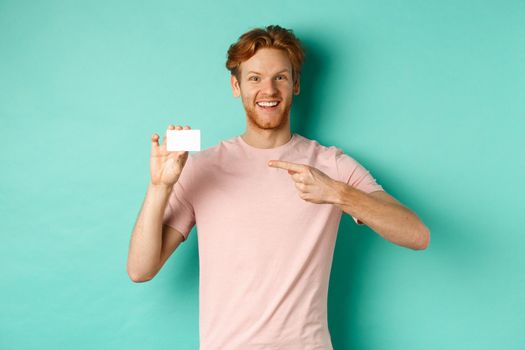 Attractive adult man with beard and red hair pointing finger at plastic credit card, smiling pleased at camera, standing over turquoise background.
