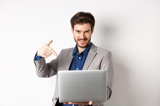 Confident young male entrepreneur working online with laptop, pointing at computer and smiling, standing on white background.