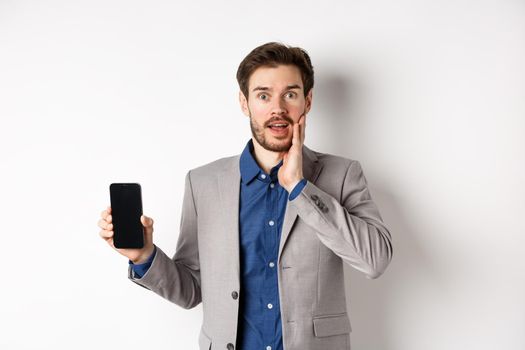 Excited business man in suit showing empty smartphone screen and gasping amazed, demonstrate application, standing on white background.