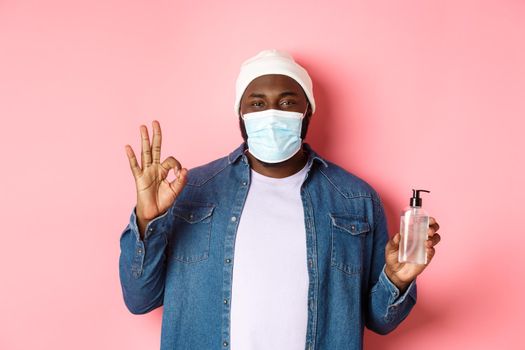 Covid-19, lifestyle and lockdown concept. Handsome african-american hipster guy in face mask and beanie, showing hand sanitizer and okay sign, advice to use antiseptic.