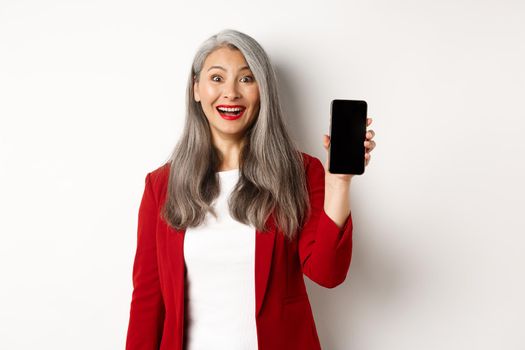 Cheerful asian female entrepreneur showing mobile phone screen, smiling amused, standing over white background.
