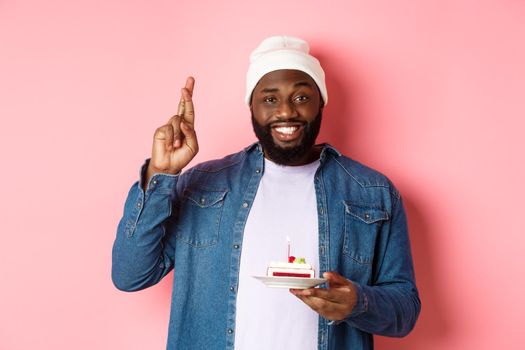 Handsome african-american guy celebrating birthday, making wish with fingers crossed, holding bday cake with candle, standing against pink background.