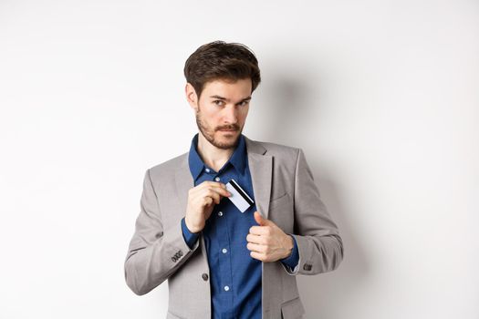 Confident and successful businessman looking self-assured while put plastic credit card in suit pocket, standing on white background.