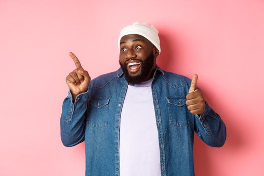 Happy african-american man showing thumb-up and pointing at upper left corner copy space, praising good deal, standing over pink background.