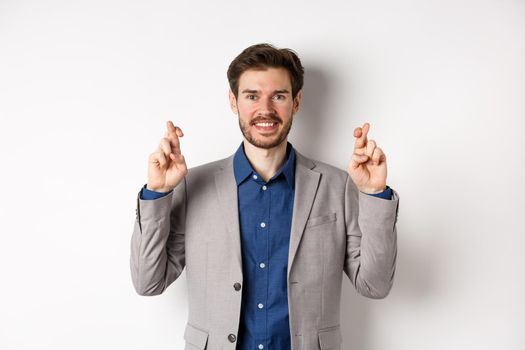 Hopeful smiling businessman looking optimistic, cross fingers good luck, praying to win, waiting for results with hope, standing on white background.