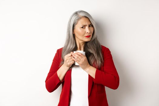 Business people concept. Greedy asian businesswoman protecting her coffee and frowning at camera, standing over white background.
