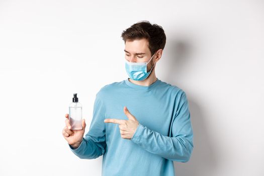 Covid-19, health and quarantine concept. Young man pointing and looking at hand sanitizer bottle, showing good antiseptic, white background.