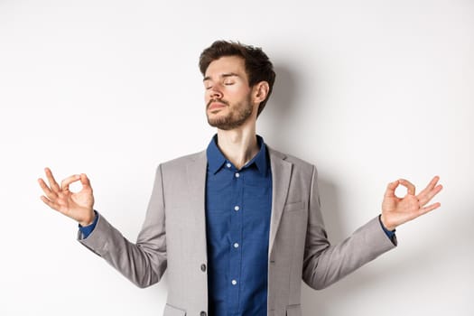 Calm and focused businessman meditating with eyes closed and hands spread sideways, finding peace in meditation, practice yoga breathing, standing on white background.