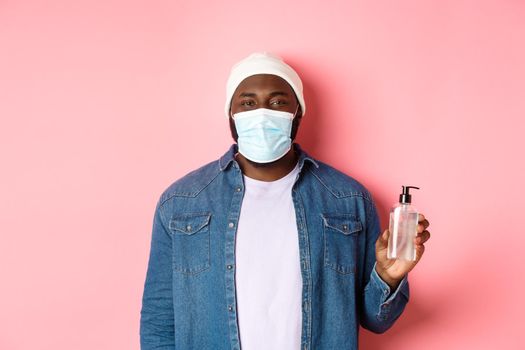 Covid-19, lifestyle and lockdown concept. Handsome hipster guy in face mask showing hand sanitizer, using antiseptic, standing over pink background.