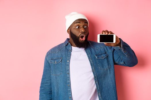 Online shopping and technology concept. Impressed african-american man in beanie showing phone screen, staring at mobile amazed, standing over pink background.