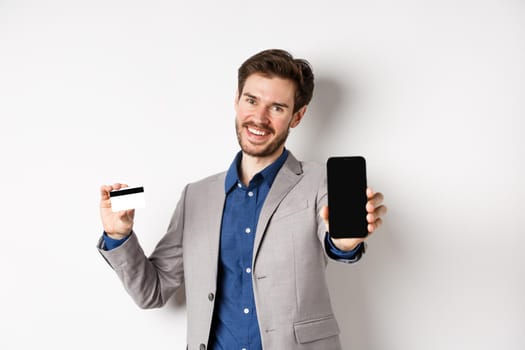 Online shopping. Successful business man in suit plastic credit card and empty cellphone screen, smiling pleased at camera, white background.