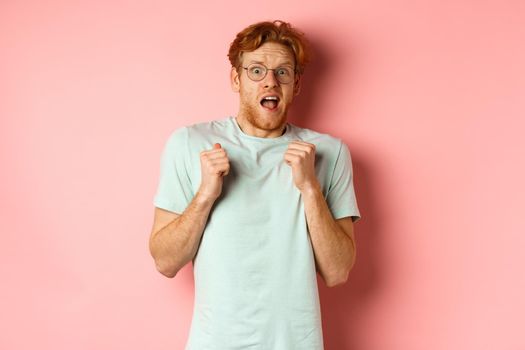 Portrait of scared redhead guy in glasses staring startled at camera, press hands to body and scream of fear, standing over pink background.