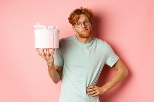 Love and holidays concept. Funny redhead guy shrugging silly and showing pink gift box for valentines day, standing over pink background.