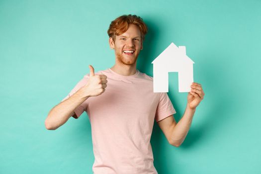 Real estate concept. Young man with red hair, wearing t-shirt, showing paper house cutout and thumb up, recommend agency, standing over turquoise background.