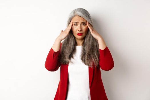 Troubled asian female entrepreneur having headache, touching head and frowning, standing over white background.