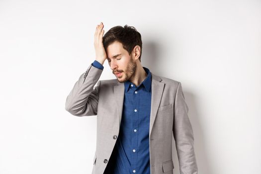 Annoyed businessman in suit facepalm with eyes closed, bothered by someone stupid, pissed-off at work, standing on white background.