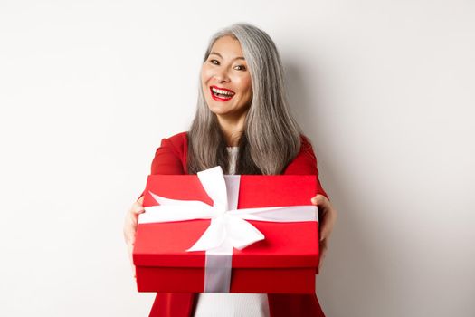 Happy holidays concept. Cheerful asian senior woman giving you red gift box and smiling, congratulating with valentine day, standing over white background.