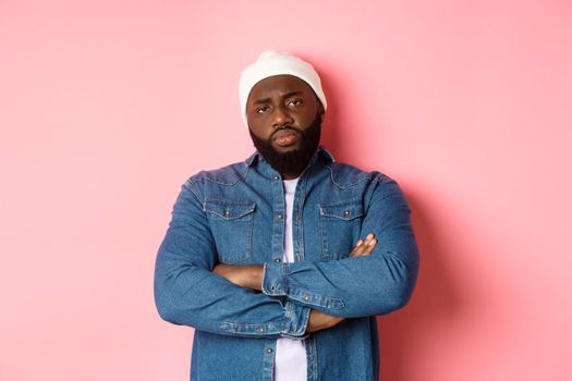 Disappointed african-american man in hipster beanie, staring at camera displeased, cross arms on chest, standing over pink background.