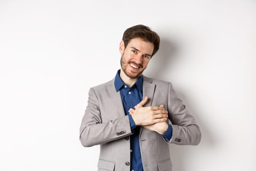 Happy man in suit holding hands on heart, feeling touched or grateful, having tender memories in soul, standing against white background.