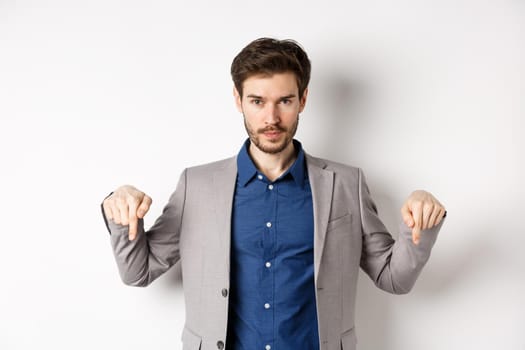 Look here. Handsome bearded man in suit pointing fingers down, looking confident at camera, showing banner, standing on white background.