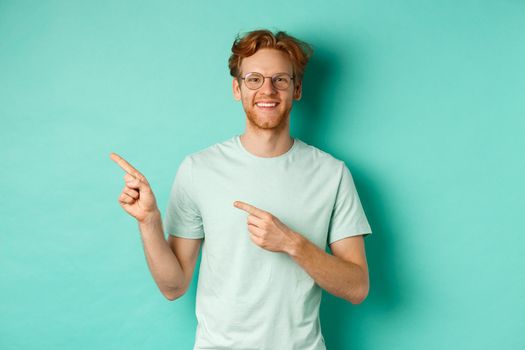 Young cheerful man with short red hair and glasses, pointing fingers left at copy space, smiling white teeth, showing advertisement, mint background.