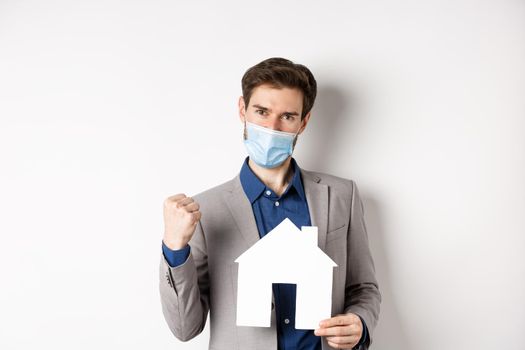 Real estate and covid-19 concept. Excited man in medical mask and suit motivated to buy house, holding paper home cutout and say yes, white background.
