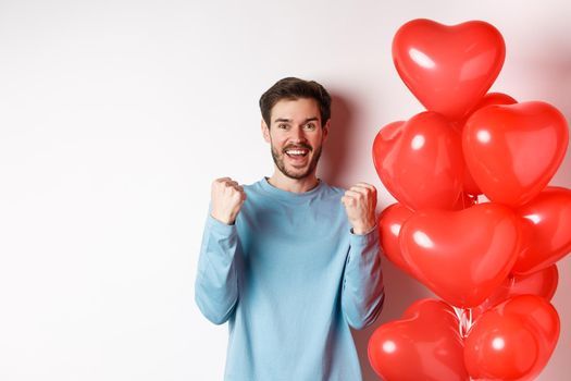Portrait of happy guy celebratiing lovers day, standing near valentines red heart balloon and cheering, making yes gesture and smiling at camera, standing over white background.